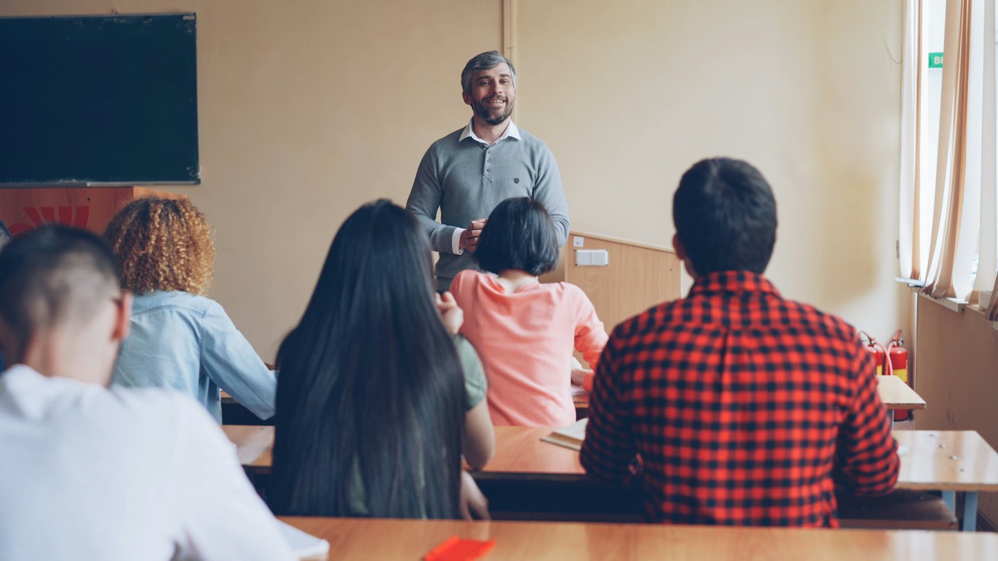 A smiling teacher addresses students in a classroom.