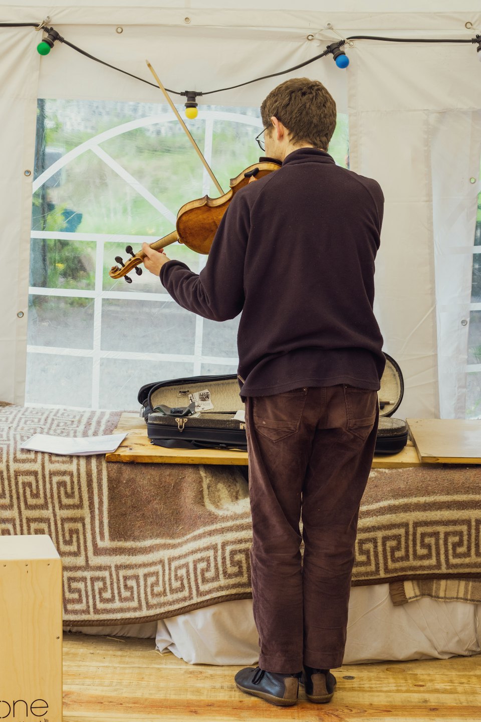 Person playing violin inside a tent with foliage visible outside.